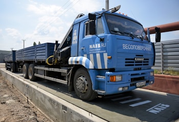 A large blue truck is parked on a weighing platform. The truck has multiple wheels and a flatbed trailer. It features text and logos on the side, and there is a metal fence in the background along with a clear sky.