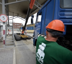 A construction site scene with a worker in an orange safety vest and helmet operating heavy machinery. A large blue truck is parked with an extended mechanical arm lifting cylindrical objects. Another person wearing a green shirt and an orange hard hat is positioned near the truck. Nearby buildings and a sheltered area can be seen in the background.