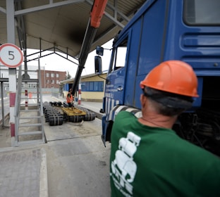A construction site scene with a worker in an orange safety vest and helmet operating heavy machinery. A large blue truck is parked with an extended mechanical arm lifting cylindrical objects. Another person wearing a green shirt and an orange hard hat is positioned near the truck. Nearby buildings and a sheltered area can be seen in the background.