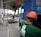 A construction site scene with a worker in an orange safety vest and helmet operating heavy machinery. A large blue truck is parked with an extended mechanical arm lifting cylindrical objects. Another person wearing a green shirt and an orange hard hat is positioned near the truck. Nearby buildings and a sheltered area can be seen in the background.