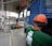 A construction site scene with a worker in an orange safety vest and helmet operating heavy machinery. A large blue truck is parked with an extended mechanical arm lifting cylindrical objects. Another person wearing a green shirt and an orange hard hat is positioned near the truck. Nearby buildings and a sheltered area can be seen in the background.