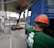 A construction site scene with a worker in an orange safety vest and helmet operating heavy machinery. A large blue truck is parked with an extended mechanical arm lifting cylindrical objects. Another person wearing a green shirt and an orange hard hat is positioned near the truck. Nearby buildings and a sheltered area can be seen in the background.