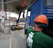 A construction site scene with a worker in an orange safety vest and helmet operating heavy machinery. A large blue truck is parked with an extended mechanical arm lifting cylindrical objects. Another person wearing a green shirt and an orange hard hat is positioned near the truck. Nearby buildings and a sheltered area can be seen in the background.