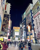 A bustling urban scene featuring a busy street lined with tall buildings covered in colorful advertisements and billboards, primarily for anime and electronics. People walk along the street, some in groups, others alone, under the bright lights of the city at night.