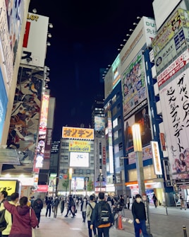 A bustling urban scene featuring a busy street lined with tall buildings covered in colorful advertisements and billboards, primarily for anime and electronics. People walk along the street, some in groups, others alone, under the bright lights of the city at night.