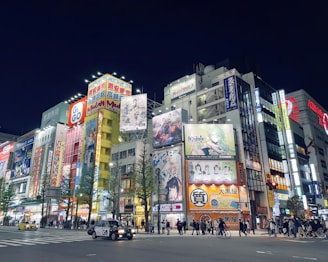 A vibrant urban street scene at night, filled with brightly lit billboards and advertisements showcasing colorful anime and video game characters. The scene includes numerous pedestrians walking along the sidewalks and some cars driving through the intersection. Tall buildings are adorned with eye-catching signage and neon lights.