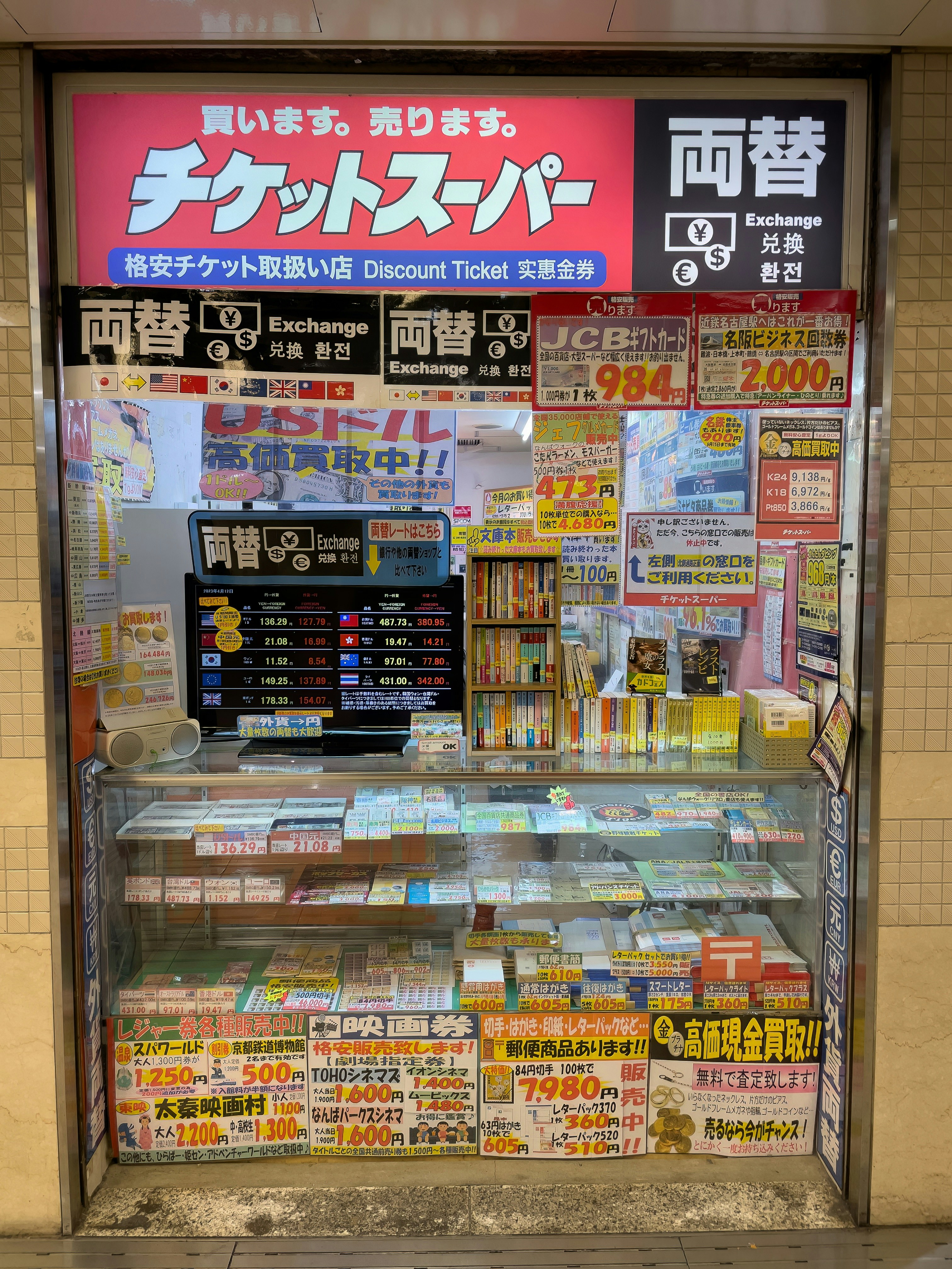 A storefront with various promotional banners and signs in multiple languages advertising ticket exchange services. The counter displays numerous tickets and small items, while the walls are covered with colorful posters and price listings.