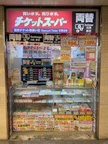 A storefront with various promotional banners and signs in multiple languages advertising ticket exchange services. The counter displays numerous tickets and small items, while the walls are covered with colorful posters and price listings.