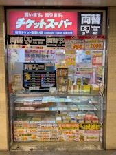 A storefront with various promotional banners and signs in multiple languages advertising ticket exchange services. The counter displays numerous tickets and small items, while the walls are covered with colorful posters and price listings.