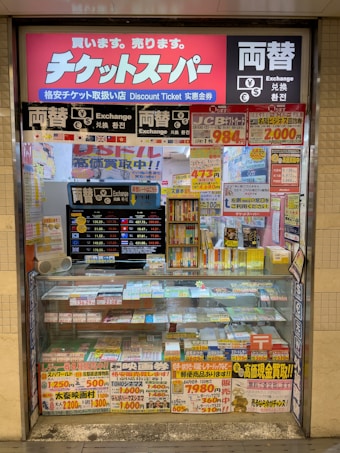 A storefront with various promotional banners and signs in multiple languages advertising ticket exchange services. The counter displays numerous tickets and small items, while the walls are covered with colorful posters and price listings.