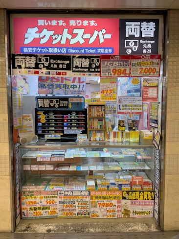 A storefront with various promotional banners and signs in multiple languages advertising ticket exchange services. The counter displays numerous tickets and small items, while the walls are covered with colorful posters and price listings.