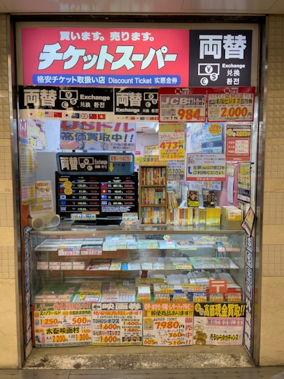 A storefront with various promotional banners and signs in multiple languages advertising ticket exchange services. The counter displays numerous tickets and small items, while the walls are covered with colorful posters and price listings.
