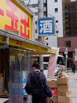 A person wearing a mask and a black backpack stands in front of a shop entrance with a yellow and red awning. The shop has signs with large printed characters and is surrounded by boxes and packaging. A sign above displays the character for alcohol. The street scene includes modern buildings and a bus.