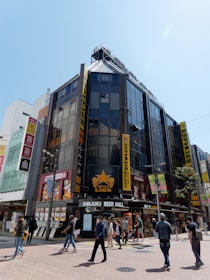 A bustling street scene featuring a large multi-story building with glass panels and various colorful signs. The building houses multiple businesses, including 'Ginza Lion,' a beer hall located on the ground floor. Several pedestrians are walking by on the sidewalk under a clear blue sky.
