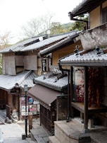 Traditional Japanese architecture with wooden buildings and tiled roofs line a narrow street. Signage with Japanese characters adorns the storefronts. Various outdoor decorations hang near the entrance. The scene is surrounded by trees, and the atmosphere feels serene and historic.
