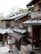 Traditional Japanese architecture with wooden buildings and tiled roofs line a narrow street. Signage with Japanese characters adorns the storefronts. Various outdoor decorations hang near the entrance. The scene is surrounded by trees, and the atmosphere feels serene and historic.