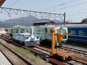 Several maintenance and service trains are positioned on railway tracks in a train yard. The scene includes a blue and white maintenance vehicle, a mint green train car with 'TOTETSU' labeling, and an orange crane or lifting equipment on the tracks. Overhead electric cables and a background view of mountains and industrial facilities contribute to the industrial setting.