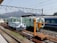 Several maintenance and service trains are positioned on railway tracks in a train yard. The scene includes a blue and white maintenance vehicle, a mint green train car with 'TOTETSU' labeling, and an orange crane or lifting equipment on the tracks. Overhead electric cables and a background view of mountains and industrial facilities contribute to the industrial setting.