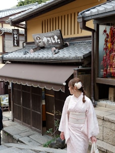 A woman dressed in a pale pink traditional kimono stands in front of a historic Japanese building with wooden details and a tiled roof. The building has a sign with Japanese characters and features hanging red ornaments to the right. The pathway is made of stone, adding to the traditional aesthetic.