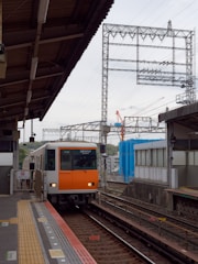 A train is approaching the platform of an outdoor train station. The train has an orange and white front. Overhead power lines and various electrical structures are visible in the background. There is some construction activity indicated by cranes in the distance. The platform is equipped with tactile paving and safety notices.