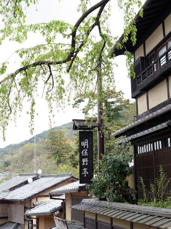 Traditional Japanese architecture with wooden structures and a black sign featuring white Japanese characters. The foreground includes a branch with green leaves, and the background consists of mountains and additional greenery.