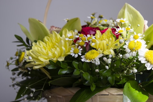 A vibrant arrangement of flowers consisting of large yellow chrysanthemums, white daisies with yellow centers, clusters of small white flowers, red roses, and lush green leaves is placed in a woven basket. The mixture of blooms creates an abundant and lively bouquet.