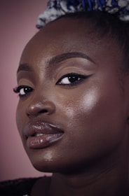 A close-up portrait of a person with smooth, glossy skin and makeup emphasizing the eyes and lips. The expression is calm and confident, with a background in soft pink.