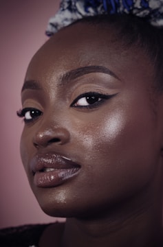 A close-up portrait of a person with smooth, glossy skin and makeup emphasizing the eyes and lips. The expression is calm and confident, with a background in soft pink.