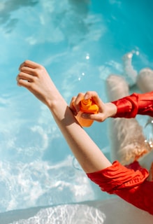 A person in a red outfit is by a pool, applying sunscreen from an orange bottle onto their arm. The clear blue water contrasts with the vibrant red of the clothing.