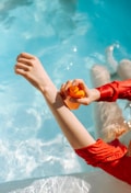 A person in a red outfit is by a pool, applying sunscreen from an orange bottle onto their arm. The clear blue water contrasts with the vibrant red of the clothing.
