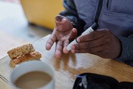 A person uses a small medical device, likely a blood glucose monitor, on their fingertip. The background includes a wooden table with two biscotti and a cup of coffee. The person is wearing a grey jacket and appears to be performing a health-related task.