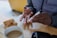 A person uses a small medical device, likely a blood glucose monitor, on their fingertip. The background includes a wooden table with two biscotti and a cup of coffee. The person is wearing a grey jacket and appears to be performing a health-related task.