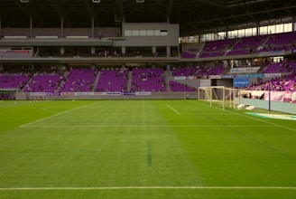 A soccer stadium with empty green grass field and goalposts. Purple seats fill the stands, and a few spectators are scattered throughout. Banners and advertisements line the balcony areas.