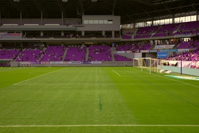 A soccer stadium with empty green grass field and goalposts. Purple seats fill the stands, and a few spectators are scattered throughout. Banners and advertisements line the balcony areas.