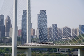 A bridge spans across a river or roadway with tall suspension cables extending vertically. In the background, several high-rise buildings form a cityscape, displaying a mix of architectural styles. The scene captures urban infrastructure and modernity.