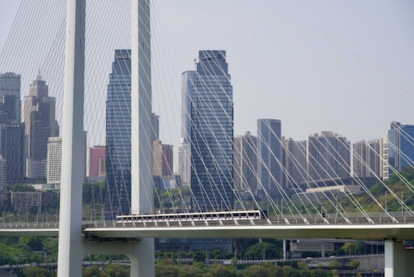 A bridge spans across a river or roadway with tall suspension cables extending vertically. In the background, several high-rise buildings form a cityscape, displaying a mix of architectural styles. The scene captures urban infrastructure and modernity.