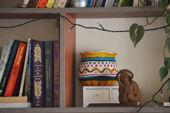 A wooden bookshelf holds a variety of books with colorful spines, including titles like 'Harry Potter' and 'The Bible'. Next to the books, there is a vibrantly painted pot with geometric patterns in red, blue, yellow, and black. Beside the pot, a small clay figurine of a person is situated. A string of fairy lights is draped across the shelf, and a leafy plant trails along the side.