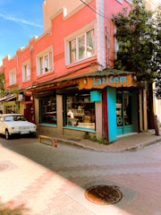 A vibrant street scene featuring a corner building painted in shades of pink and orange, with a rustic-looking shop front displaying the name 'Latifem' in turquoise letters. The shop has large glass windows, through which a person can be seen inside. A white vintage car is parked on the cobblestone street in front. The scene is enhanced by a clear blue sky and lush greenery growing on the side of the building.