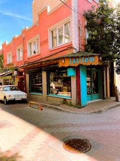 A vibrant street scene featuring a corner building painted in shades of pink and orange, with a rustic-looking shop front displaying the name 'Latifem' in turquoise letters. The shop has large glass windows, through which a person can be seen inside. A white vintage car is parked on the cobblestone street in front. The scene is enhanced by a clear blue sky and lush greenery growing on the side of the building.