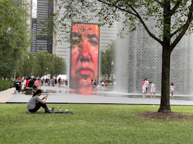 A large outdoor installation featuring a tall, digital screen displaying a human face with water cascading around it. Several people are nearby, some sitting and others walking around. A tree provides shade over a grassy area, where a person is sitting on the ground.