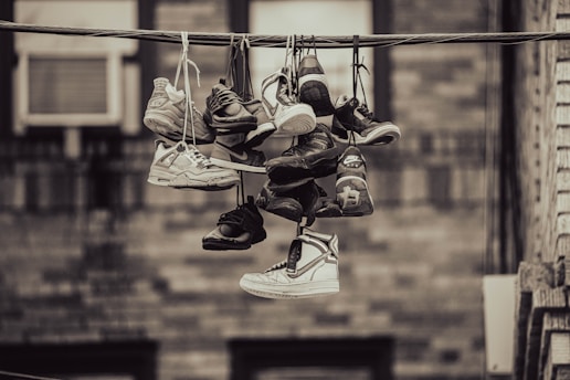 A collection of various sneakers are hanging from a telephone wire by their laces. The sneakers vary in style and color, suggesting a vibrant array of footwear. The background features an out-of-focus wall, giving the scene an urban feel.