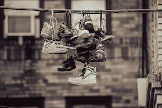 A collection of various sneakers are hanging from a telephone wire by their laces. The sneakers vary in style and color, suggesting a vibrant array of footwear. The background features an out-of-focus wall, giving the scene an urban feel.