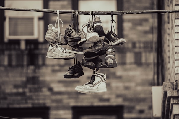 A collection of various sneakers are hanging from a telephone wire by their laces. The sneakers vary in style and color, suggesting a vibrant array of footwear. The background features an out-of-focus wall, giving the scene an urban feel.