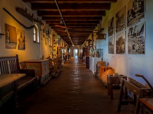 A long corridor in a museum is lined with various artifacts and artworks displayed on both sides. The walls feature black-and-white photographs and vintage farming tools. There are wooden benches and large wooden chests with intricate designs. The overhead wooden beams add to the rustic atmosphere, with soft lighting creating a warm atmosphere.