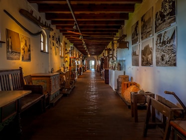A long corridor in a museum is lined with various artifacts and artworks displayed on both sides. The walls feature black-and-white photographs and vintage farming tools. There are wooden benches and large wooden chests with intricate designs. The overhead wooden beams add to the rustic atmosphere, with soft lighting creating a warm atmosphere.