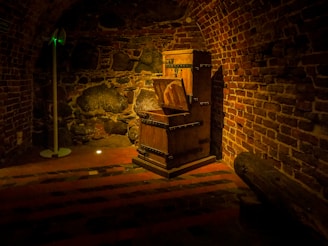 A dimly lit underground brick chamber with a wooden medieval torture device, possibly an Iron Maiden, prominently placed against the stone wall. The atmosphere is eerie and rustic, with a solitary spotlight highlighting the wooden structure. The curved ceiling and stone wall suggest a dungeon-like setting, while shadows cast by the single light add to the mysterious ambiance.