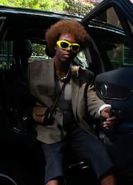 A person with a distinctive afro hairstyle is wearing bright yellow sunglasses and casual yet stylish attire, including a checkered blazer, mesh top, and shorts. They are seated inside a car with the door open, holding the car door handle.
