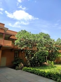 A well-maintained garden area with lush green trees and bushes alongside a building with a tiled roof and multiple windows. The sky is clear with a few fluffy white clouds, adding a serene aspect to the environment.