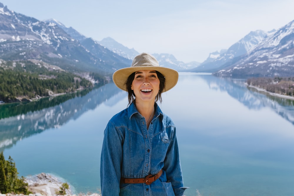 a woman wearing a hat standing in front of a lake