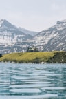 A large building resembling a lodge or hotel is situated on a grassy hill overlooking a calm, expansive body of water. Snow-capped mountains rise in the background under a pale blue sky.